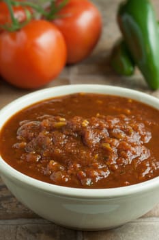 Fresh salsa in a white bowl on a tile surface with tomatoes and jalapenos in the background.