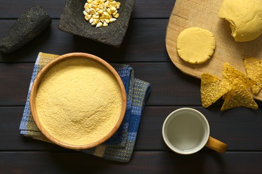 Overhead shot of cornmeal in wooden bowl with corn kernels in mortar, cup of water and cornmeal dough on wooden board, photographed on dark wood with natural light