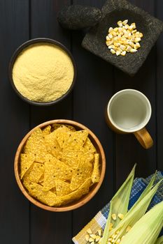 Overhead shot of tortilla chips in wooden bowl surrounded by its ingredients water, cornmeal and corn, photographed on dark wood with natural light