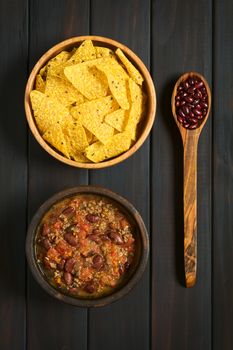 Overhead shot of chili con carne and tortilla chips in bowls with dried kidney beans on wooden spoon, photographed on dark wood with natural light