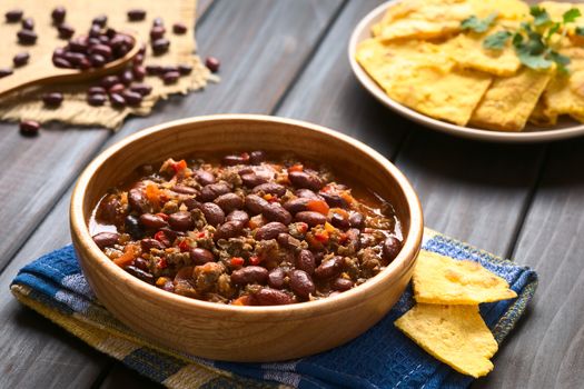 Wooden bowl of chili con carne with homemade tortilla chips in the back, photographed with natural light (Selective Focus, Focus in the middle of the chili)