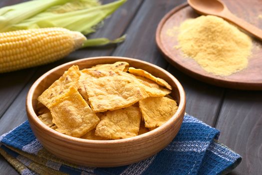 Wooden bowl of homemade baked corn chips with cobs of corn and cornmeal in the back, photographed with natural light (Selective Focus, Focus on the front of the chip on the top)