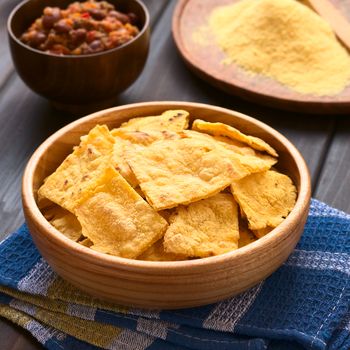 Wooden bowl of homemade baked corn chips with cornmeal and a bowl of chili con carne in the back, photographed with natural light (Selective Focus, Focus on the front of the chip on the top)