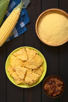 Overhead shot of homemade baked corn chips on plate with cornmeal, corn cobs and a small bowl of chili con carne, photographed on dark wood with natural light