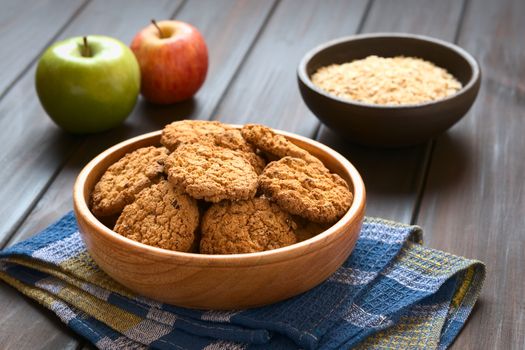 Oatmeal and apple cookies in wooden bowl with apples and oatmeal in the back, photographed with natural light (Selective Focus, Focus on the front of the cookie in the middle)