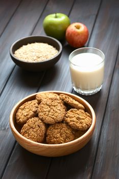 Oatmeal and apple cookies in wooden bowl with a glass of milk, apples and oatmeal in the back, photographed with natural light (Selective Focus, Focus on the front of the cookie in the middle)