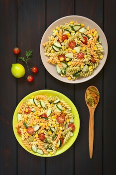 Overhead shot of two plates of vegetarian pasta salad made of tricolor fusilli, sweet corn, cucumber and cherry tomato, photographed on dark wood with natural light