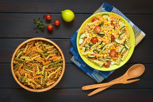 Overhead shot of raw fusilli pasta in wooden bowl and a plate of vegetarian pasta salad made of tricolor fusilli, sweet corn, cucumber and cherry tomato, photographed on dark wood with natural light