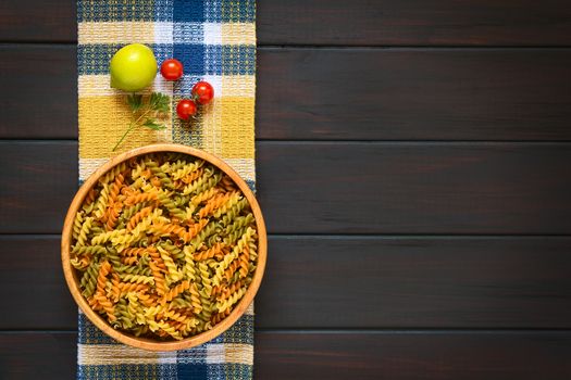 Overhead shot of raw tricolor fusilli or rotini pasta (traditional, tomato and spinach taste) in wooden bowl with ingredients, photographed on dark wood with natural light