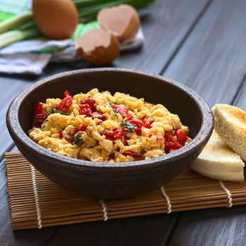 Scrambled eggs made with red bell pepper and green onion in rustic bowl with toasted bread on the side and eggs in the back, photographed with natural light (Selective Focus, Focus one third into the eggs) 