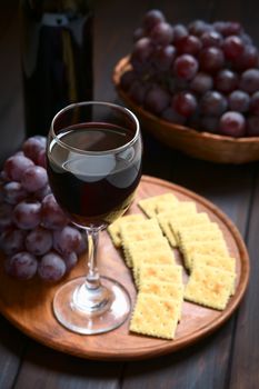 Glass of red wine with red globe grape, crackers and a bottle of wine, photographed on dark wood with natural light (Selective Focus, Focus on the front rim of the wine glass)