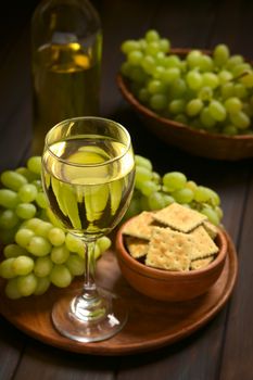 Glass of white wine with white grape, crackers and a bottle of wine, photographed on dark wood with natural light (Selective Focus, Focus on the front rim of the wine glass)