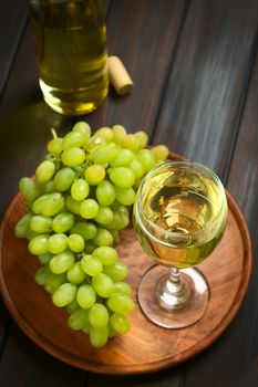 Glass of white wine with white grapes and a bottle of wine, photographed on dark wood with natural light (Selective Focus, Focus on the front rim of the wine glass)