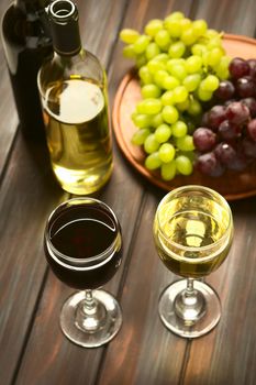 Glasses of red and white wine with red globe and white grapes and bottles of wine in the back, photographed on dark wood with natural light (Selective Focus, Focus on the front rim of the wine glasses)