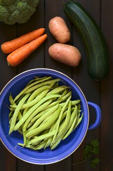 Overhead shot of green beans in blue metal strainer with parsley, broccoli, carrot, potato and zucchini, photographed on dark wood with natural light