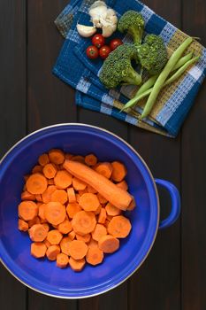 Overhead shot of fresh raw sliced carrot in blue metal strainer with broccoli, cherry tomato, garlic, green bean on kitchen towel, photographed on dark wood with natural light