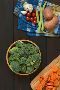 Overhead shot of fresh raw broccoli florets in wooden bowl with carrot slices on wooden board, potato, green bean, cherry tomato and garlic on kitchen towel, photographed on dark wood with natural light
