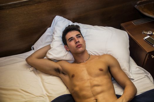 Handsome shirtless athletic young man laying in bed at night looking at the ceiling, seen from above