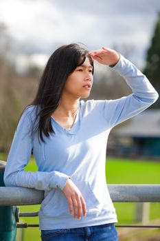 Young biracial teen girl standing, leaning against railing at park shading eyes to look off to side