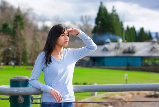 Young biracial teen girl standing, leaning against railing at park shading eyes to look off to side
