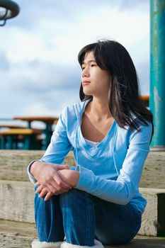 Young biracial teen girl in blue shirt and jeans sitting on wooden steps outdoors on overcast cloudy day