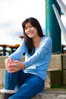 Young biracial teen girl in blue shirt and jeans sitting on wooden steps outdoors on overcast cloudy day