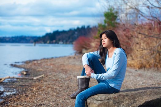 Young biracial teen girl in blue shirt and jeans sitting along rocky lake shore on bright overcast day outdoors