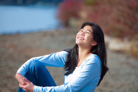 Young biracial teen girl in blue shirt and jeans sitting along rocky lake shore on bright overcast day outdoors