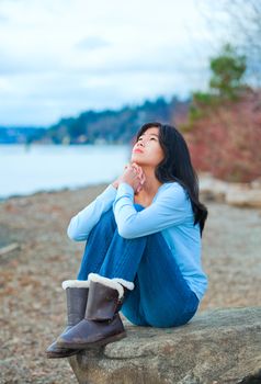 Young biracial teen girl in blue shirt and jeans sitting on boulder or rock  along lake shore praying, face upward to sky