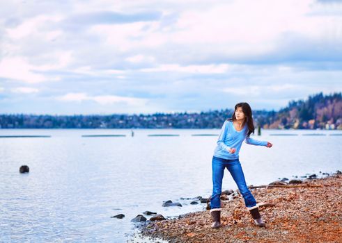 Young biracial teen girl in blue shirt and jeans along rocky lake shore, throwing rocks into the water on cloudy overcast day