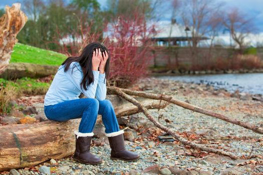 Sad young biracial teen girl in blue shirt and jeans sitting on log along rocky beach by lake
