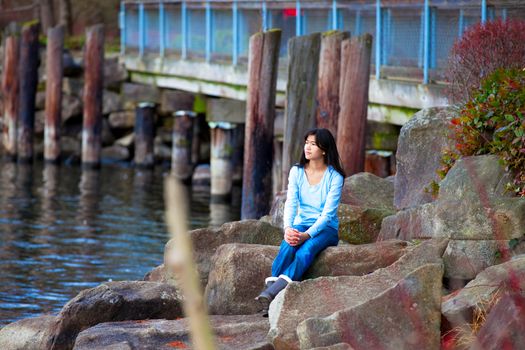 Young biracial teen girl in blue shirt and jeans sitting on large boulders along lake shore, looking out over water