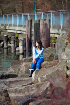 Young biracial teen girl in blue shirt and jeans sitting on large boulders along lake shore, looking out over water