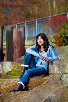 Young biracial teen girl in blue shirt and jeans sitting on large boulder, smiling and reclining