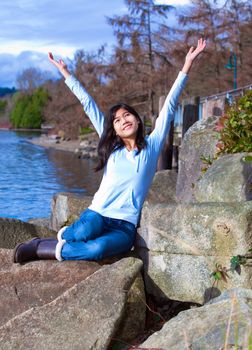 Happy young biracial teen in blue shirt and jeans sitting on large rock by lake shore, arms raised in praise.