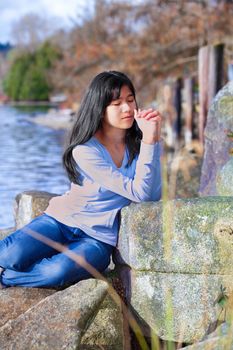 Young biracial teen girl in blue shirt and jeans quietly sitting outdoors leaning on rocks praying