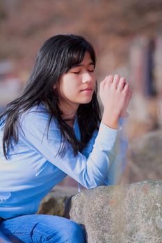 Young biracial teen girl in blue shirt and jeans quietly sitting outdoors leaning on rocks praying