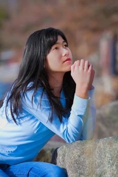 Young biracial teen girl in blue shirt and jeans quietly sitting outdoors leaning on rocks praying