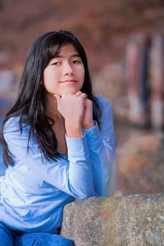 Young teen biracial girl in blue shirt and jeans quietly resting elbows on rock along lake shore, sitting  with serious expression