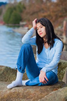 Young unhappy biracial teen girl in blue shirt and jeans sitting on rocks along lake shore, looking off to side, resting head in hand and one knee raised.