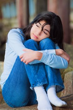 Sad biracial teen girl in blue shirt and jeans sitting on rocks along lake shore with knees pulled up to chest, lonely expression