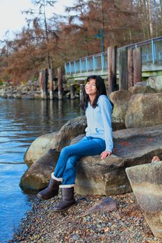 Young biracial teen girl in blue shirt and jeans sitting on large boulder or rocks along rocky lake shore, smiling and reclining