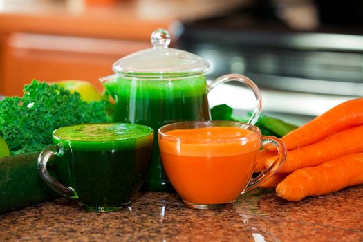 Two cups of fresh vegetable juice on kitchen counter with vegetables in background. Raw carrots, kale, spinach, cucumber, apples.