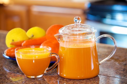 One cup of orange colored juice on kitchen counter with fruit and vegetables in background. Carrots, apples, lemons, oranges.