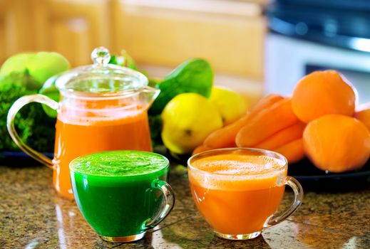 Two cups of fresh vegetable juice on kitchen counter with vegetables in background. Kale, spinach, cucumber, carrots, apples, lemons, lime, oranges.