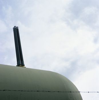 A barbed wire fence encloses a large green industrial tank with chimney 