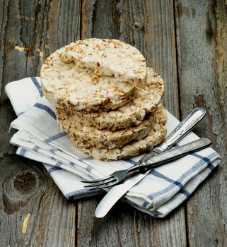 Stack of Puffed Spelt Oat Galettes with Silverware Fork and Knife on Checkered Napkin closeup on Rustic Wooden background. Health Eating Concept