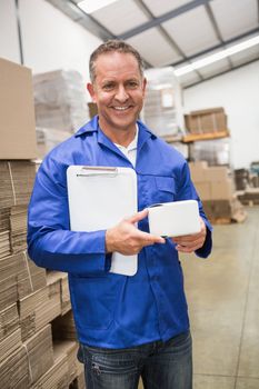 Smiling warehouse worker holding small box and clipboard in a large warehouse