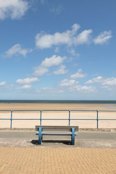 A blue painted bench and blue railings with a view across a deserted sand beach with the sea and blue cloudy sky in the distance.