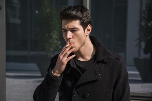 Handsome stylish young man smoking outside in urban setting, looking away